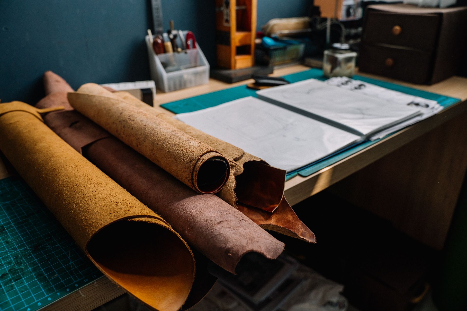 Rolls of tanned leather on a workbench beside a cutting mat.